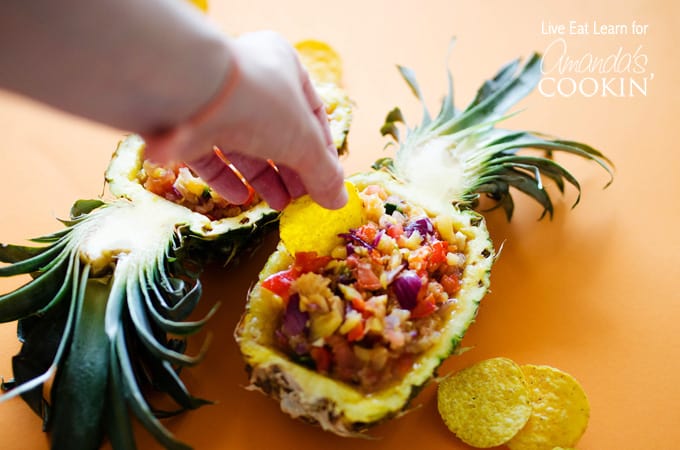 A photo of a tortilla chip scooping pineapple salsa from a halved pineapple.