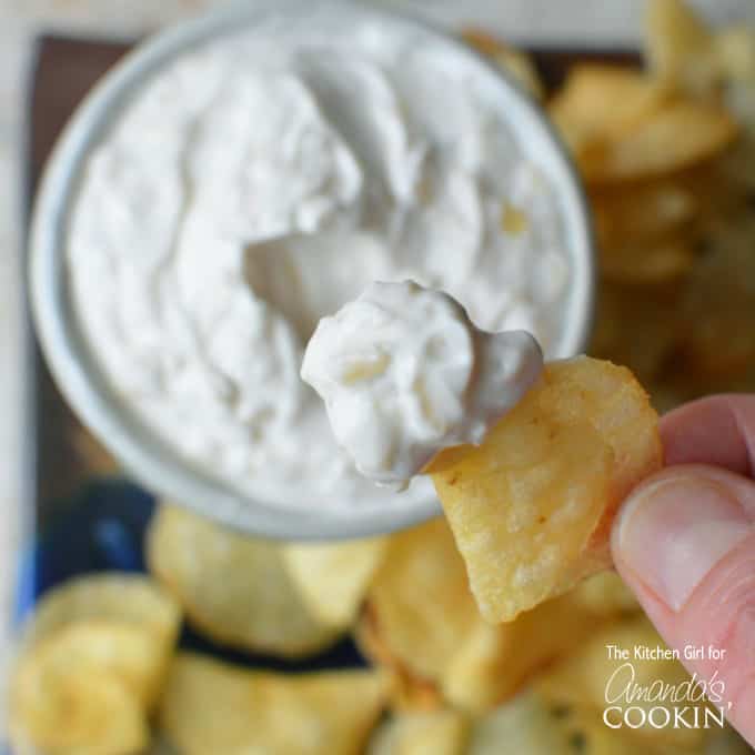 A close up of a potato chip with skinny French onion dip on it and the bowl of dip in the background.