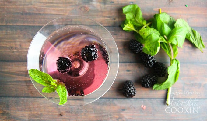 An overhead of a blackberry gin martini topped with fresh blackberries and mint leaves and some on the table next to the glass.