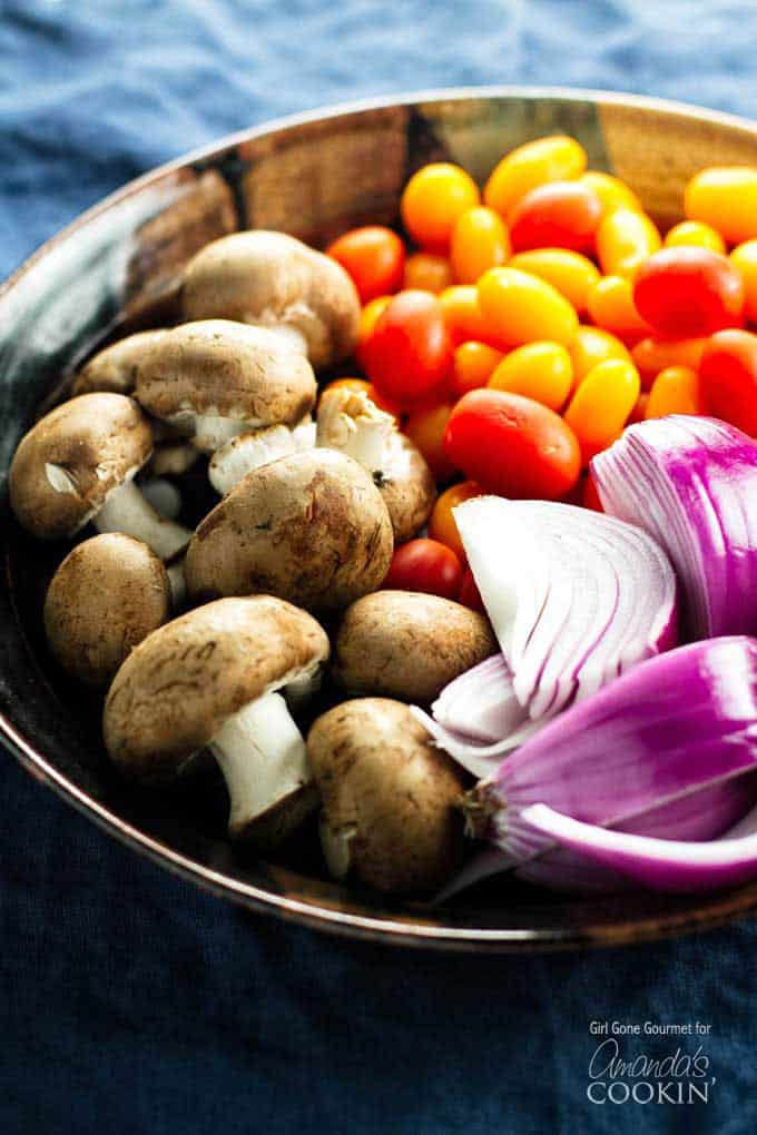 A close up photo of mushrooms, chopped red onion and tomatoes in a bowl.