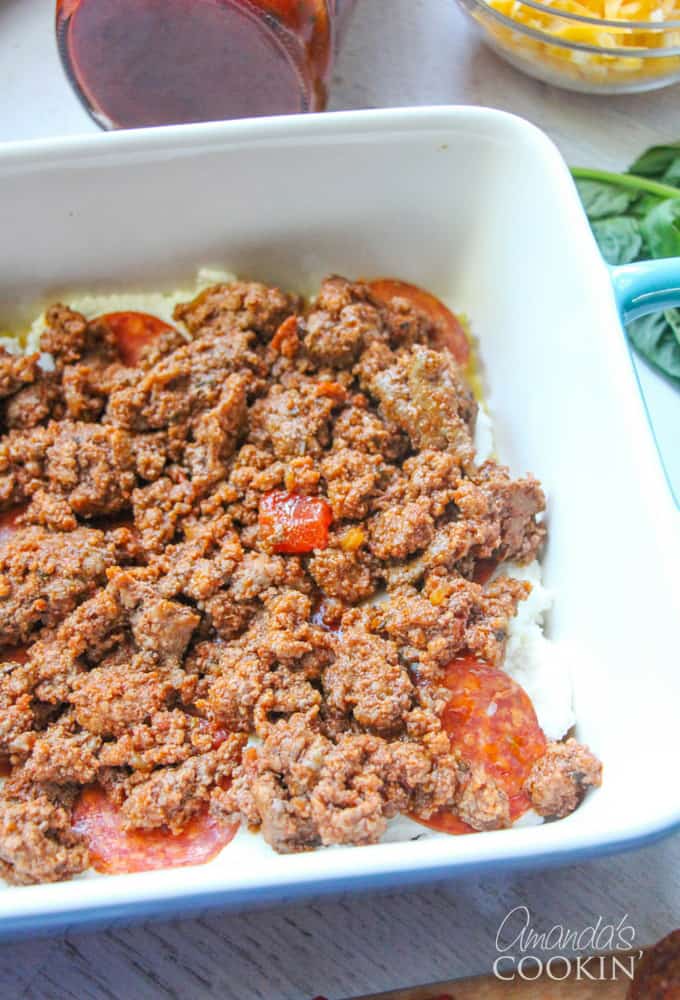 An overhead photo of cooked ground beef over pepperoni, ricotta cheese and lasagna noodles.