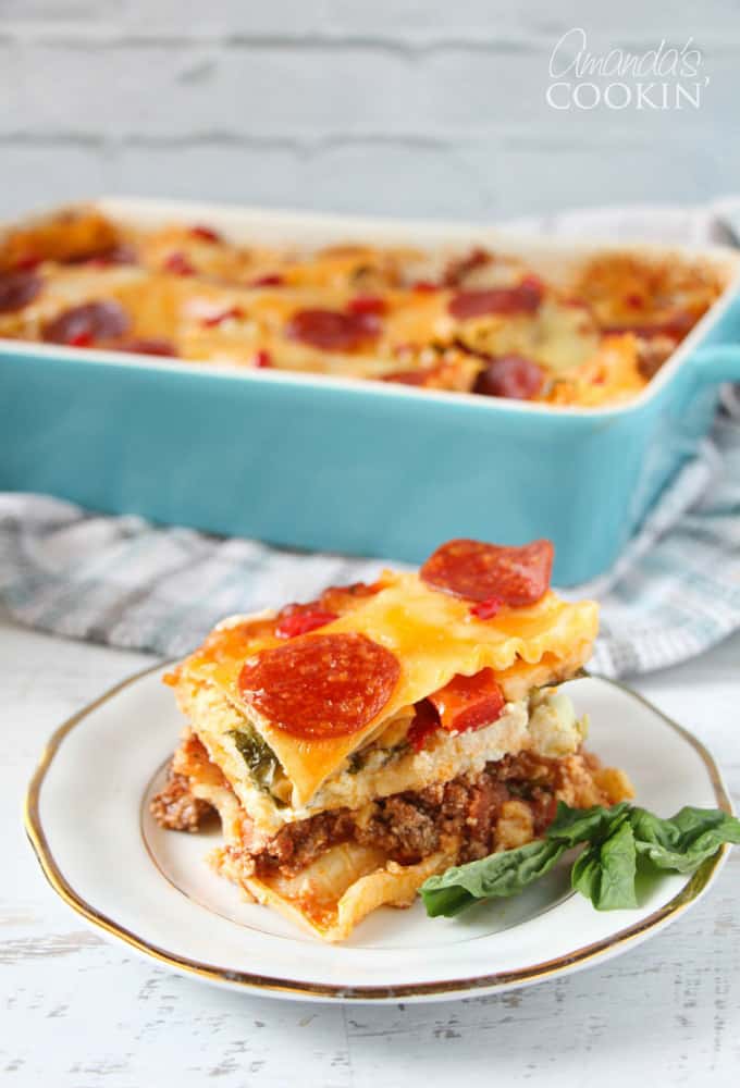 A close up photo of pizza lasagna on a white and gold plate with the casserole pan in the background. 