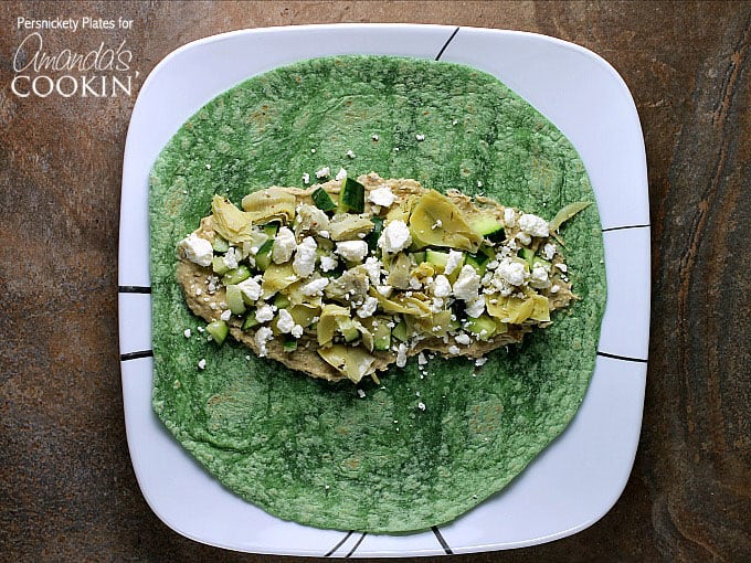 An overhead photo of chopped artichoke hearts, diced cucumber, hummus and crumbled feta on a spinach wrap.
