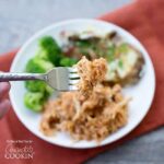 A close up of BBQ crockpot chicken on a fork with a plate of more in the background.