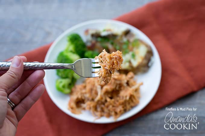 A close up of BBQ crockpot chicken on a fork with a plate of more in the background.