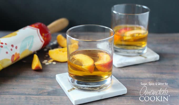 Two short clear glasses filled with peach pie cocktail with two peach slices floating on top.