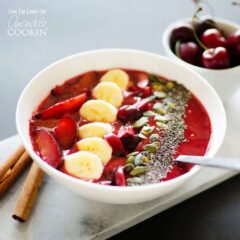 A close up photo of a stone fruit smoothie in a white bowl topped with sliced plums, bananas cherries, mixed nuts and seeds.