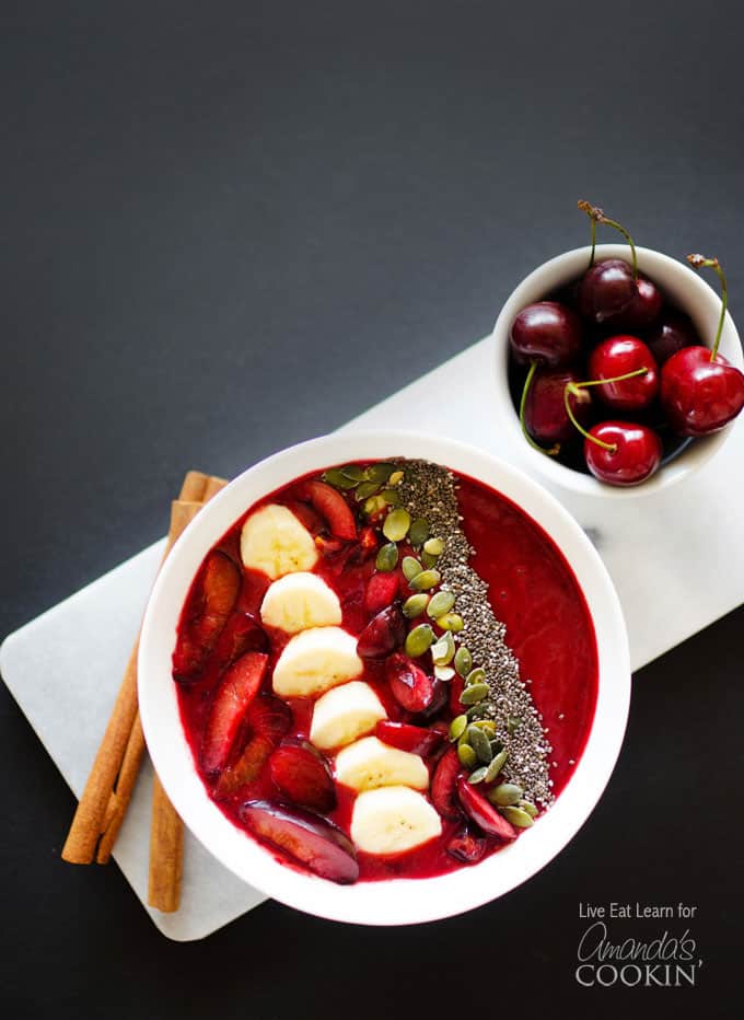 An overhead photo of a stone fruit smoothie in a white bowl topped with sliced plums, bananas cherries, mixed nuts and seeds.