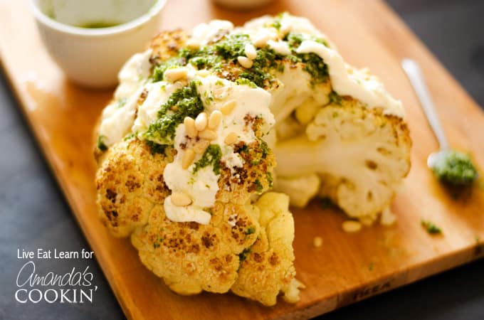 A close up of a whole roasted cauliflower topped with whipped feta and herb sauce on a wooden cutting board.