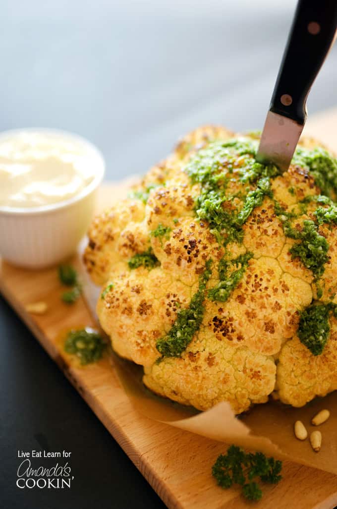 A close up of a whole roasted cauliflower topped with whipped feta and herb sauce on a wooden cutting board with a knife in the center.