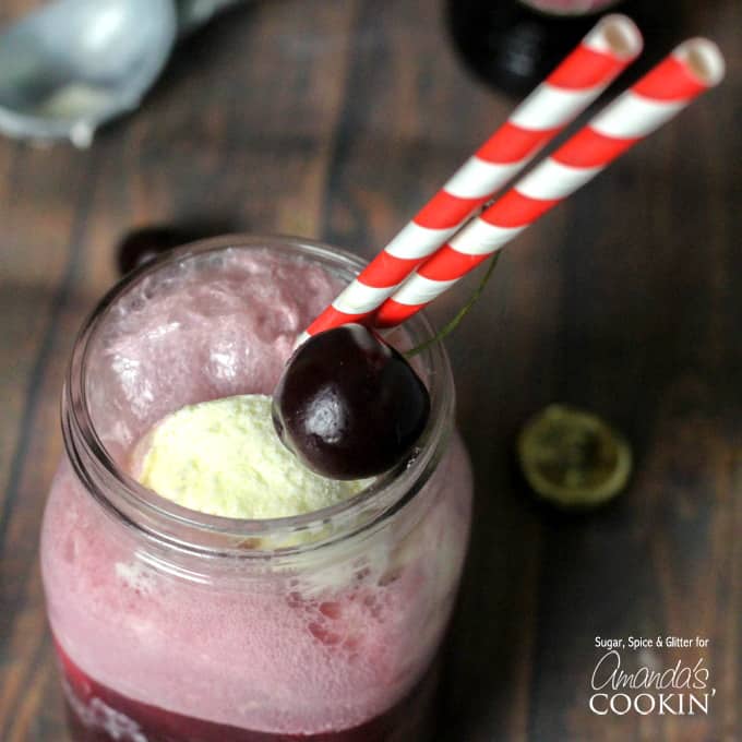 A close up of a mason jar filled with a black cherry float cocktail and served with red and white straws and a cherry.