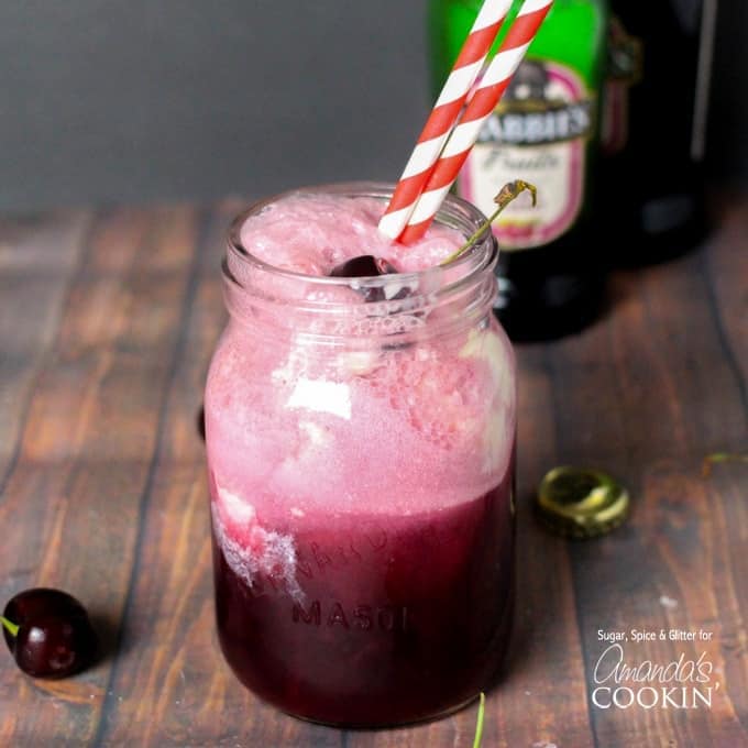 A mason jar filled with a black cherry float cocktail and served with red and white straws and topped with a cherry.