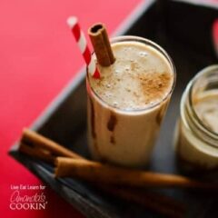 A tall clear glass filled with a cinnamon bun smoothie and served with a cinnamon stick and a red stripped straw.