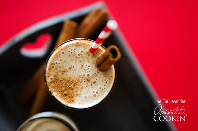 An overhead of a tall clear glass filled with a cinnamon bun smoothie and served with a cinnamon stick and a red stripped straw.