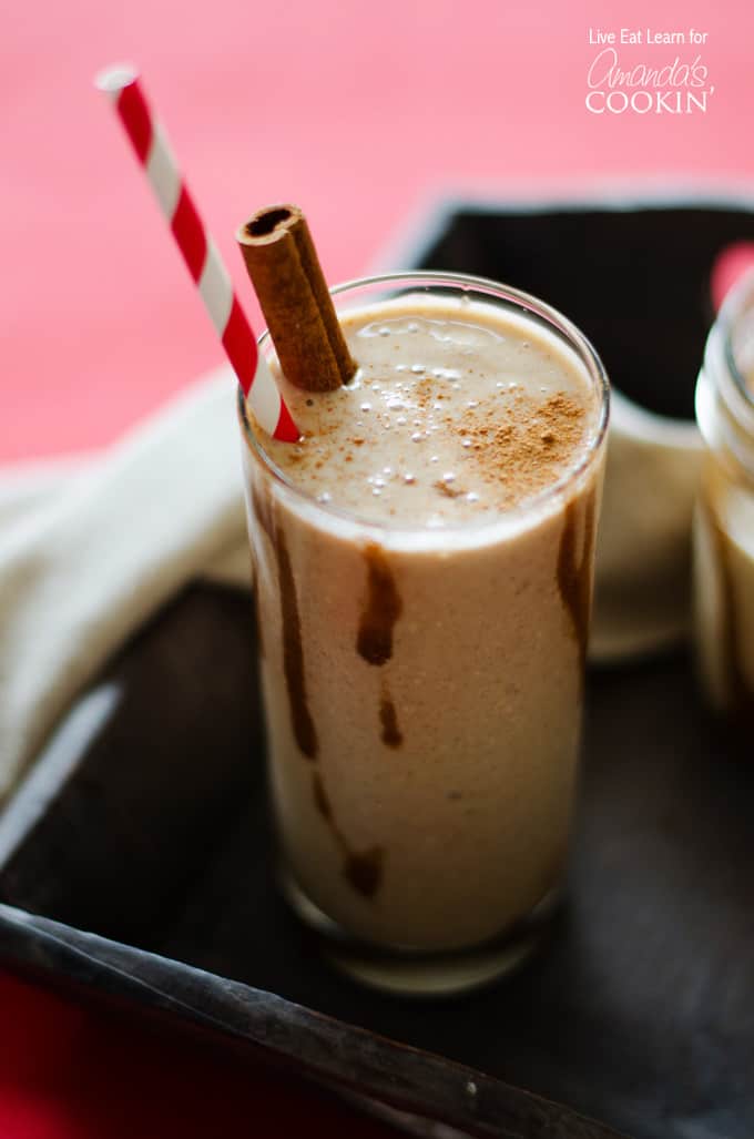 A close up of a tall clear glass filled with a cinnamon bun smoothie and served with a cinnamon stick and a red stripped straw.