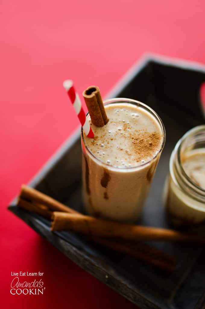 A tall clear glass filled with a cinnamon bun smoothie and served with a cinnamon stick and a red stripped straw.