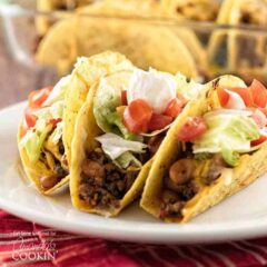 Three hard shell beef and bean oven tacos resting on a white plate topped with lettuce, tomatoes and sour cream.