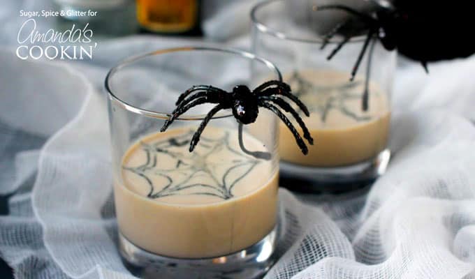 A close up of two clear glasses filled with a spiderweb in cream martini and a fake spider on the glass.