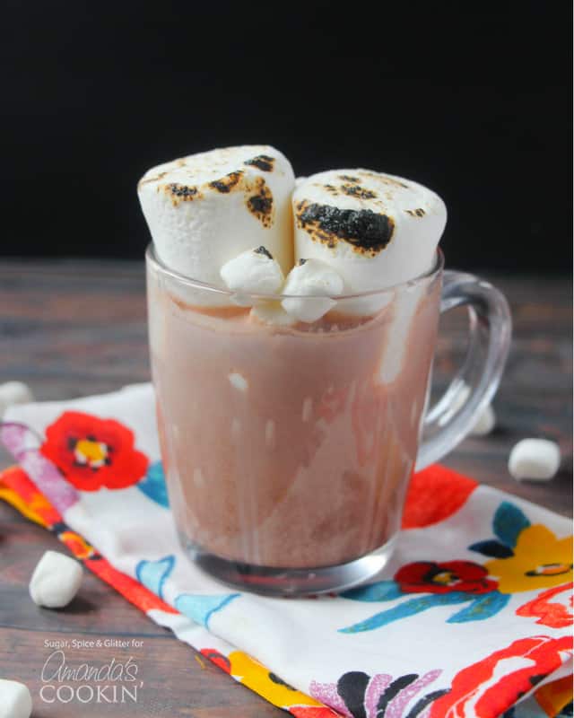 A clear mug filled with a hot chocolate cocktail and two roasted marshmallows on top.