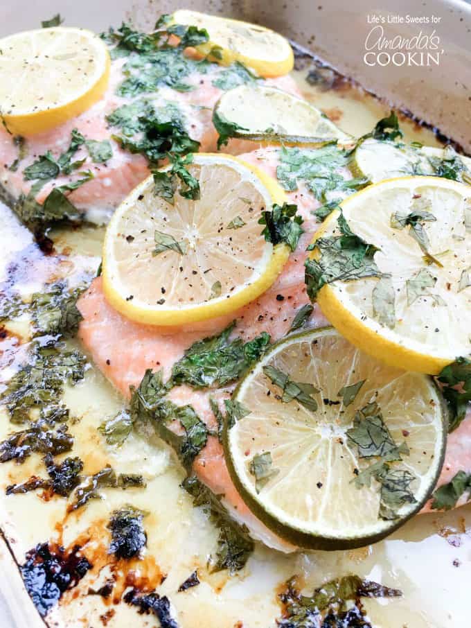 A close up photo of baked salmon topped with lemon and lime wheels, cilantro and butter.
