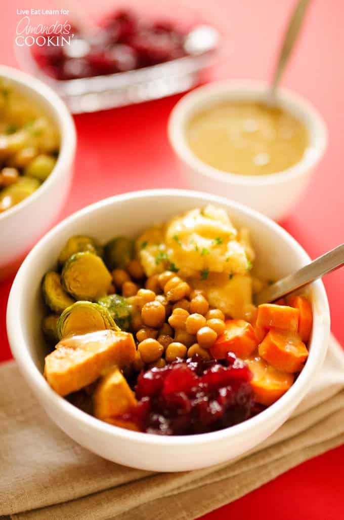 A close up of a Thanksgiving leftovers bowl filled with chickpeas, carrots, brussles sprouts, sweet potatoes, mashed potatoes, cranberry sauce and gravy.