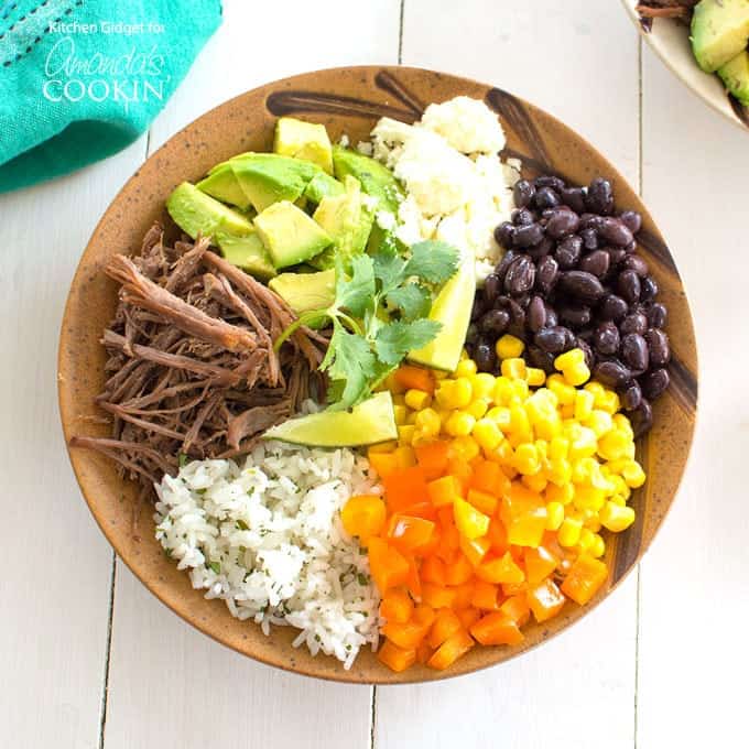 An overhead of a brown bowl filled with white rice, bell pepper, corn, black beans, crumbled queso fresco, avocado, lime and cooked beef.