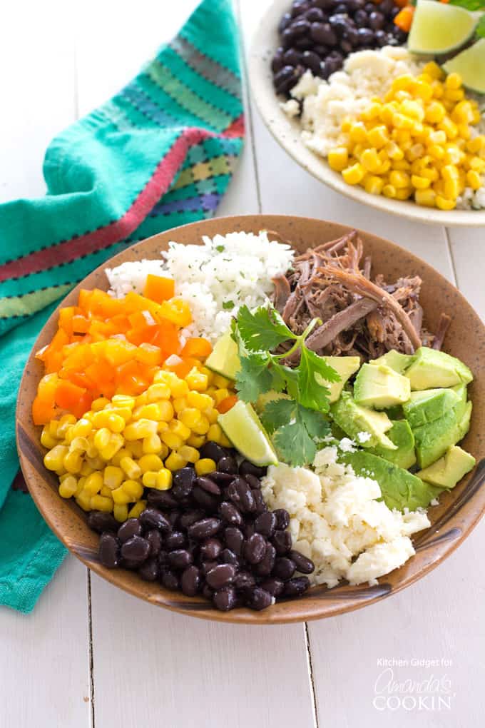 An overhead of a brown bowl filled with white rice, bell pepper, corn, black beans, crumbled queso fresco, avocado, lime and cooked beef.