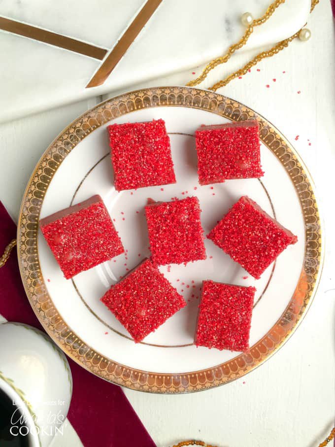 Squares of red velvet fudge with red sprinkles on top sitting on a white and gold plate.