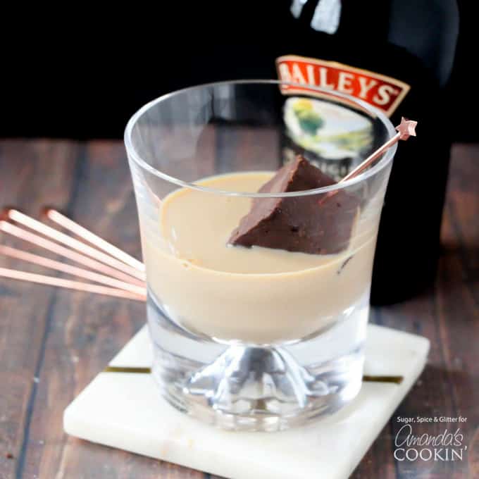 A clear glass resting on a wooden cutting board filled with Bailey\'s and a piece of fudge on a stir stick.