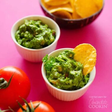 Two white bowls of guacamole with a bowl of chips in the background and two tomatoes.