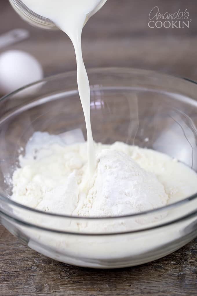 A clear glass bowl filled with flour and milk being poured in.