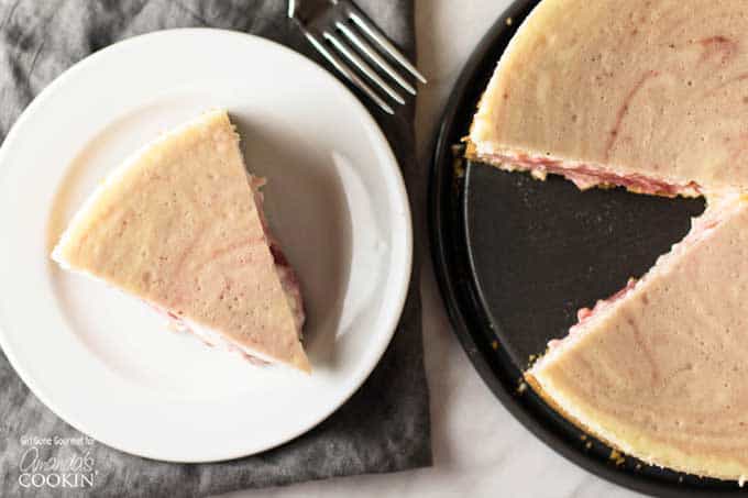 A slice of strawberry swirled cheesecake on a white plate sitting next to the entire baked cheesecake.