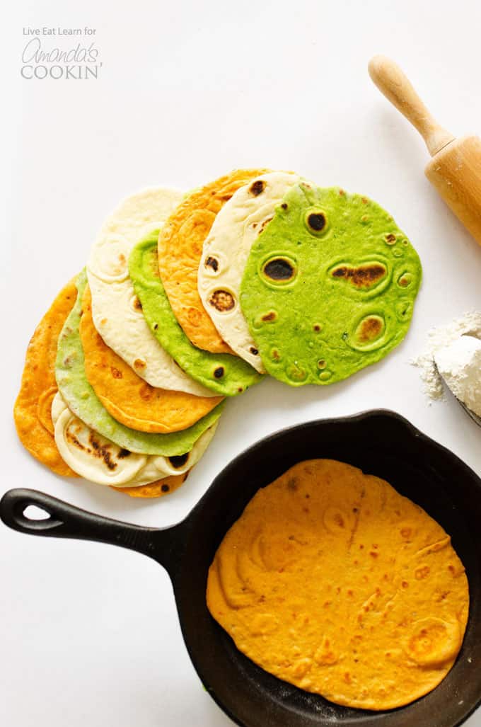 An overhead picture of a homemade tortilla resting in a skillet and multiple kinds of homemade tortillas resting on a white cutting board.