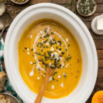 Slow cooker butternut squash soup in a white crock with a wooden spoon, topped with pepitas, coconut milk and fresh thyme