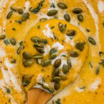 Close up of a wooden spoon in creamy butternut squash soup topped with pepitas, fresh thyme and a swirl of coconut milk