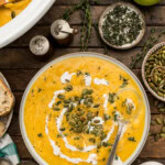Bowl of butternut squash soup with pepitas and a spoon beside the slow cooker, green apples, fresh thyme and salt and pepper shakers