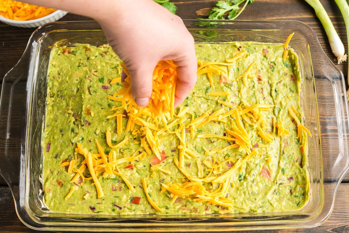 sprinkling cheddar cheese over guacamole in pan