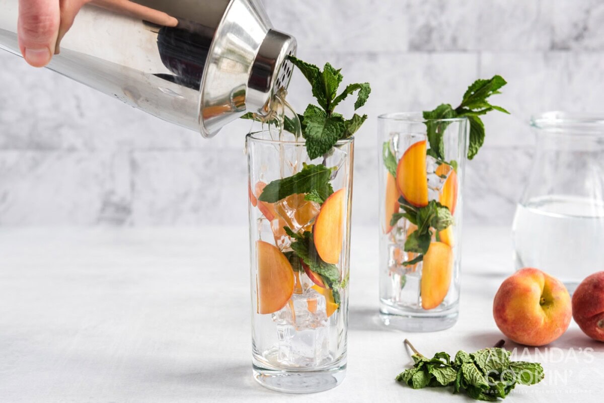 cocktail shaker pouring peach mojito into glass with peaches and mint