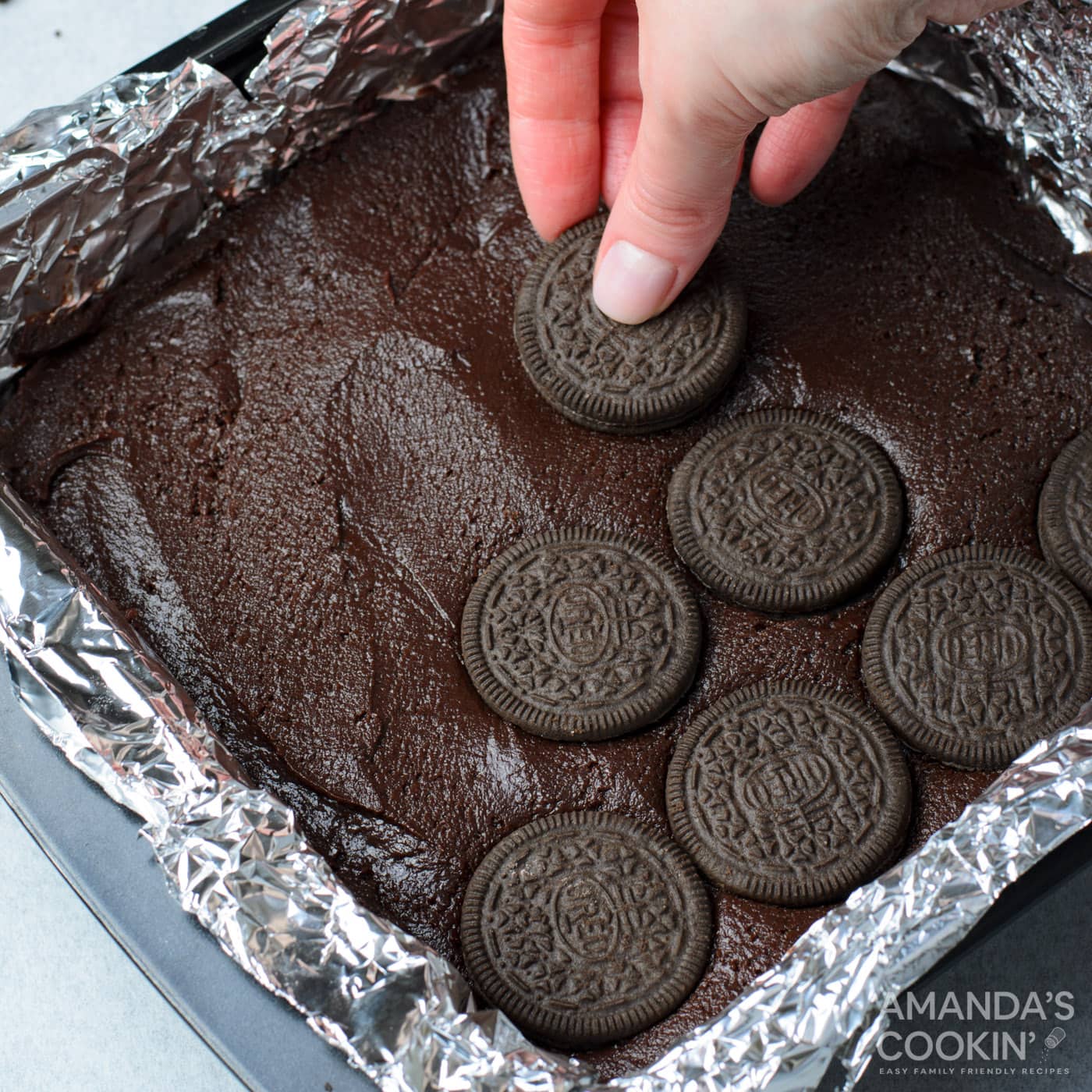 hand placing oreo cookies on top of brownie batter in a pan