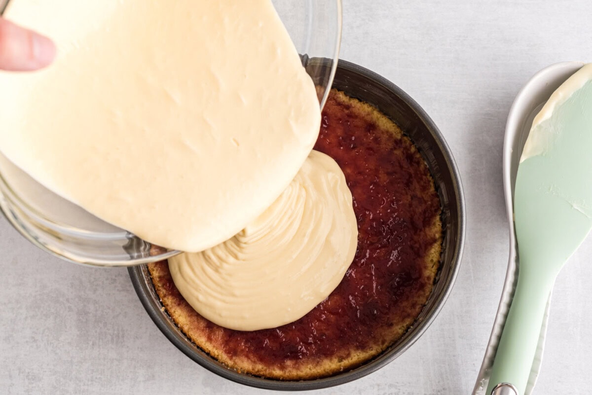 pouring cheesecake mixture on top of raspberry preserves and cake