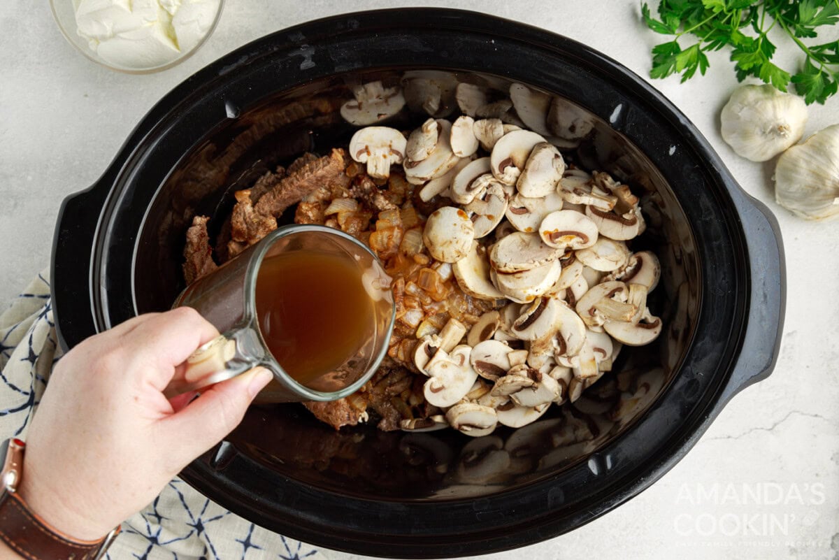 pouring beef stock into crockpot with mushrooms,, onions, and steak