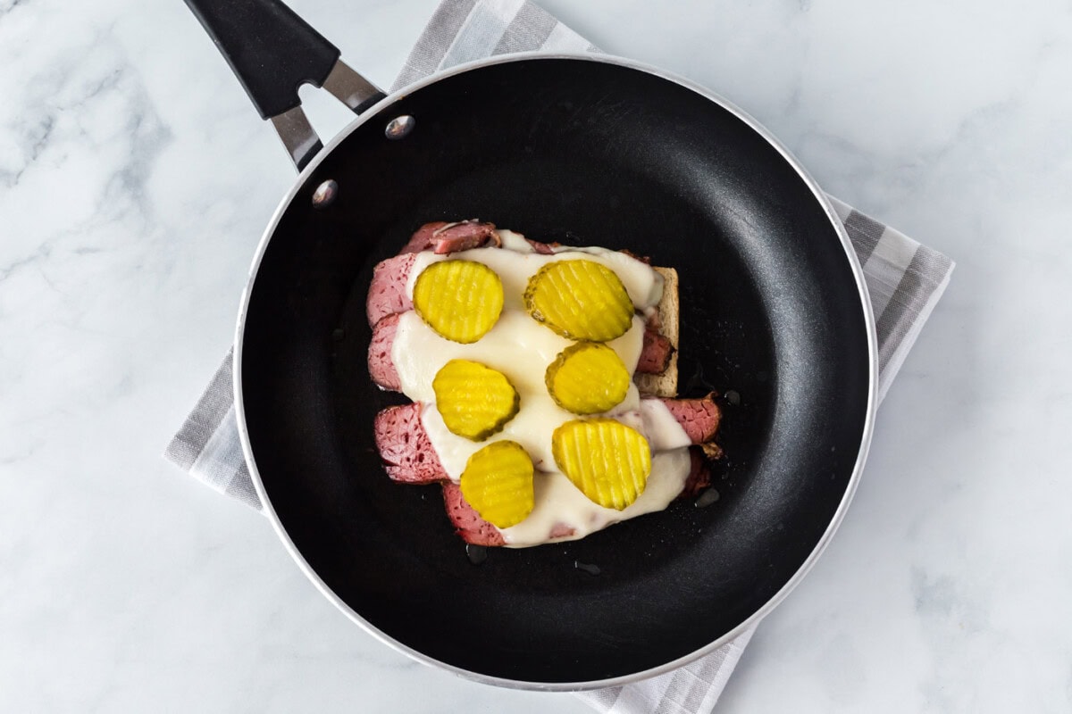 bread with corned beef, cheese, and pickle chips in a skillet