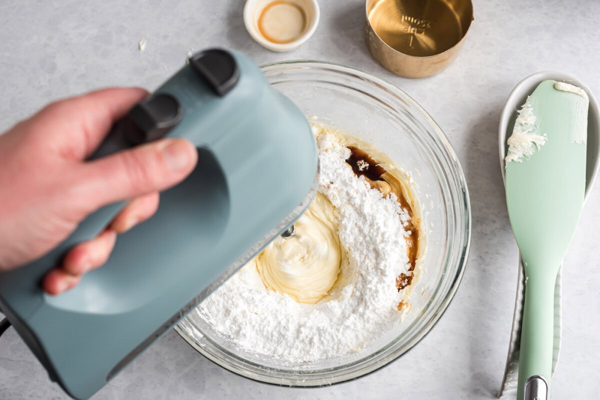 vanilla extract, cream cheese, and powdered sugar being mixed with a hand mixer