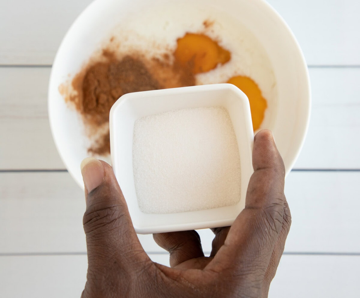 Hand holding sugar over bowl of spices and egg mixture