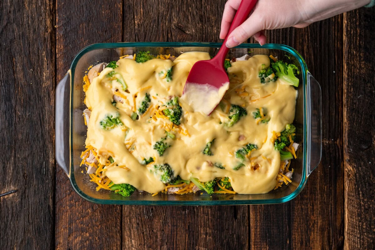 spatula spreading cream of chicken over broccoli, chicken, and cheese
