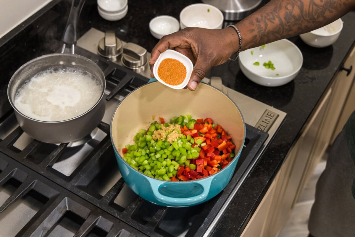 hand adding cajun seasoning to bell pepper, celery, onion, and garlic in a dutch oven