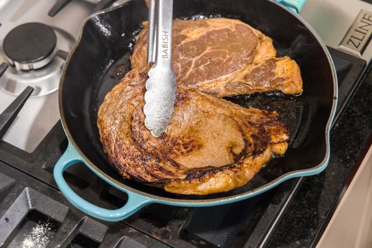 tongs flipping ribeye steak over in a skillet