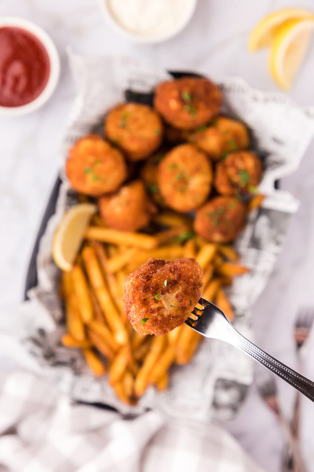 A Fried Scallop on a seafood fork held above a basket of Fried Scallops and fries