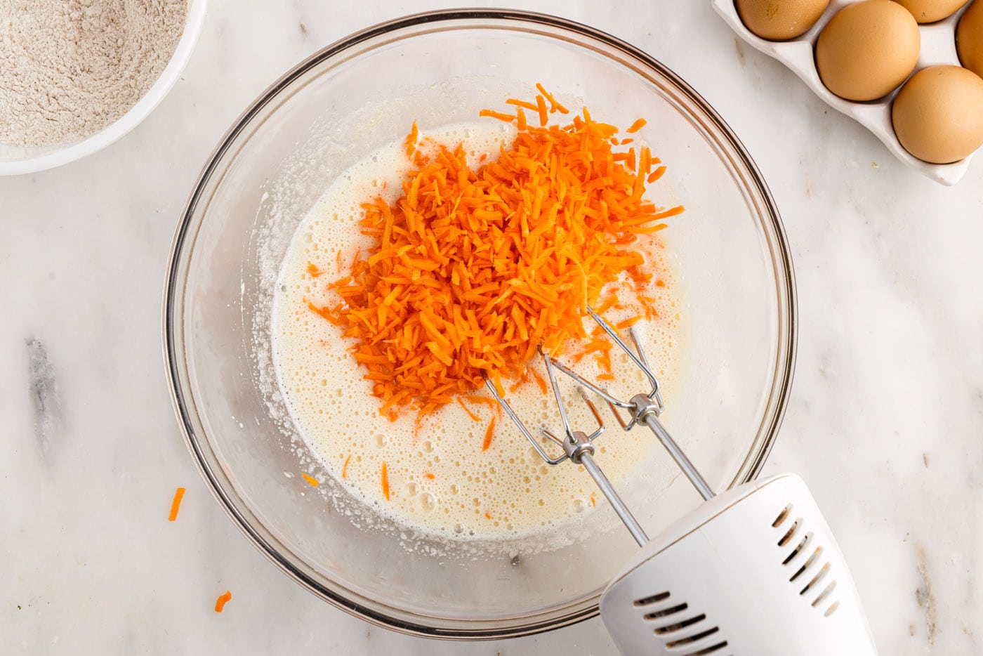 grated carrots added to egg mixture in a bowl with a mixer