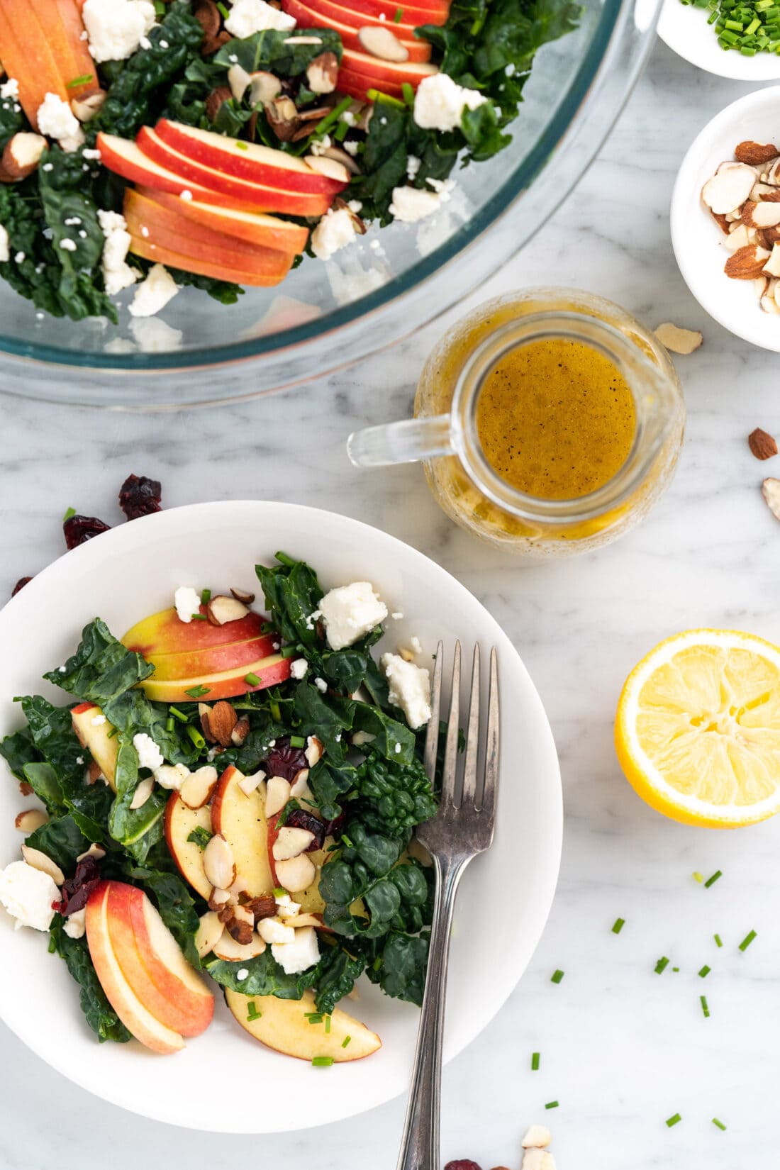 Lemon Salad Dressing in a dressing container surrounded by a plate an bowl of Kale Salad
